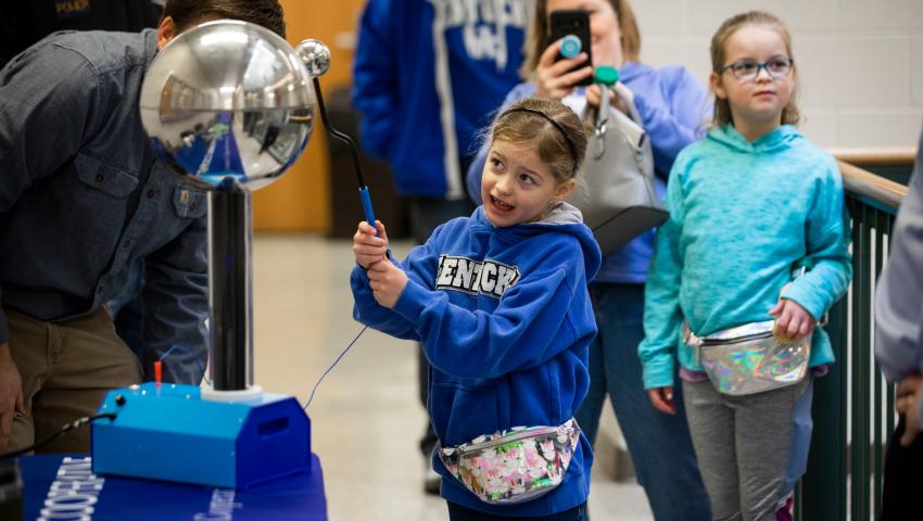 A young girl in a blue Kentucky hoodie interacts with a Van de Graaff generator, causing her hair to stand up, while people watch and take photos.